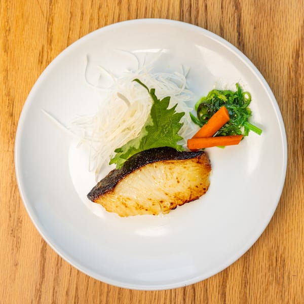 Grilled fish with shredded daikon, green shiso leaf, seaweed salad, and carrot sticks on a white plate.